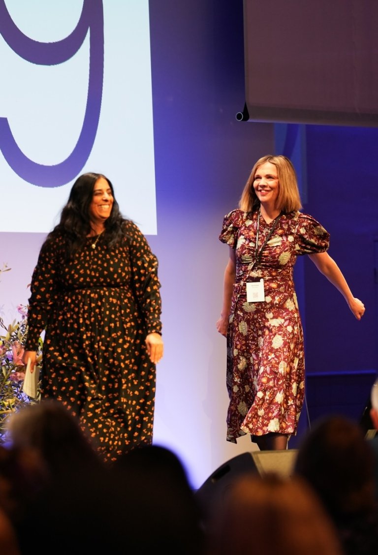 Foto av Lubna Jaffery og Inga Bolstad som går på en scene. Bak dem er det skjermer med tallet 9 på.