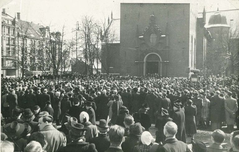 Langfredag 3. april 1942 utanfor domkyrkja i Oslo. Eit tusental menneske hadde møtt opp for å høyre gudstenesta til den avsette biskopen Eivind Berggrav. Men biskopen var nekta å forrette og arrestert av styresmaktene. Folk song ”Gud signe vårt dyre fedreland” og ”Ja vi elsker” i protest utanfor kyrkja. Hird og politi forsøkte forgjeves å spreie mengda. NTBs krigsarkiv/Ue/L0099/Livet i Norge under okkupasjonen. RA/PA-12091.