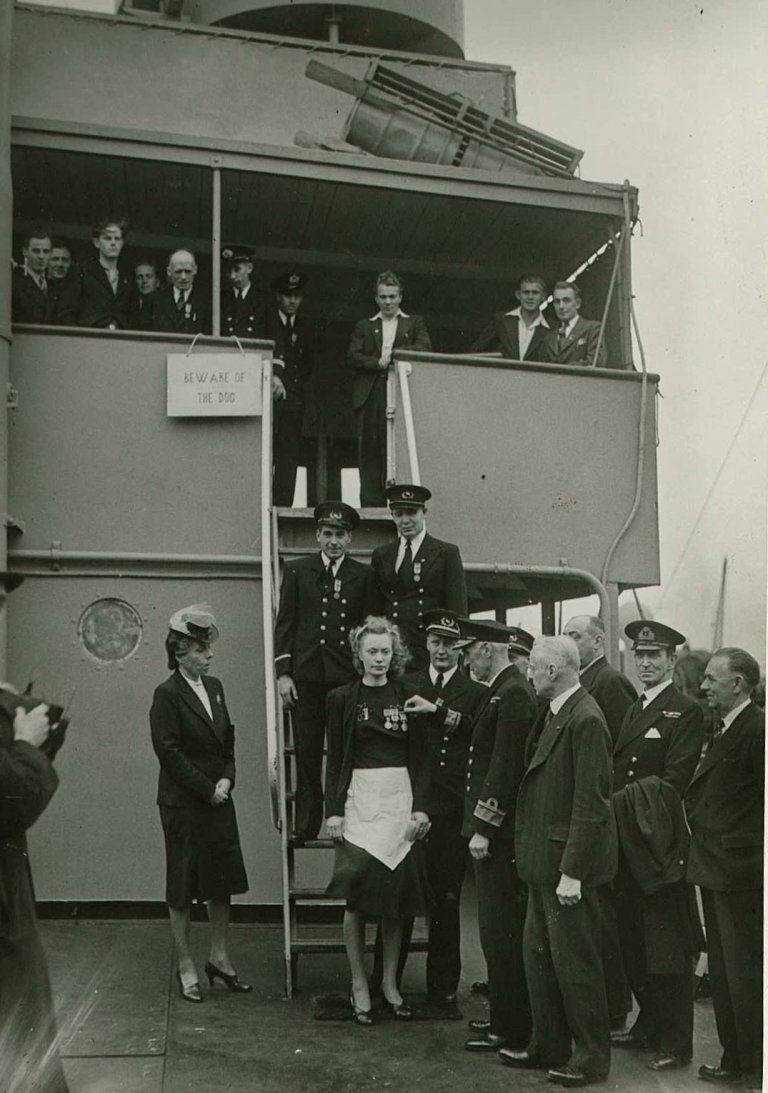 Høgtideleg dekorering av ein del av mannskapet frå M/S Talabot. Salongjenta Margit Johnsen frå Ålesund står i sentrum med serveringsforklede og tek imot British Empire Medal av admiral J.S. Ritchie. Modige Malta-Margit blei ved same høve også tildelt Krigsmedaljen og St. Olavsmedaljen med eikegrein. Ho er einaste kvinne av i alt 779 personar som har fått denne høge utmerkinga. Margit Johnsen hadde tidlegare også motteke Distinguished Service Cross på Malta. (PA-1209 NTBs krigsarkiv, Uf-123)